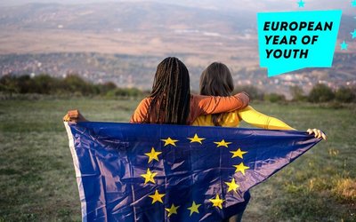 Sujetbild Europäisches Jahr der Jugend 2022: Zwei Frauen stehen mit dem Rücken zur Kamera und schauen auf eine Wiese. Gemeinsam halten sie eine Flagge mit dem EU Emblem.