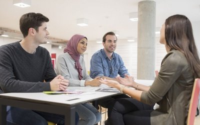 Bild von Studierenden der Wirtschaftsuniversität Wien.  Zwei Männer und zwei Frauen sitzen um einen eckigen Tisch und unterhalten sich. Eine Frau trägt ein fliederfarbiges Kopftuch, alle Studierenden lächeln.