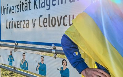 Eine Person mit ukrainischer Flagge steht neben einem Ortsschild mit dem Text "Universität Klagenfurt/Univerza v Celuvcu"