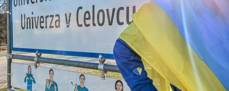 Eine Person mit ukrainischer Flagge steht neben einem Ortsschild mit dem Text "Universität Klagenfurt/Univerza v Celuvcu"