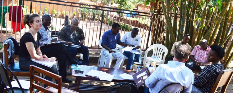 Zwei Frauen und sieben Männer sitzen rund um einen auf offener Straße stehenden Tisch in Kampala, Uganda und halten ein Meeting ab