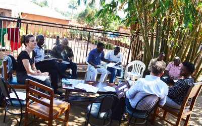 Zwei Frauen und sieben Männer sitzen rund um einen auf offener Straße stehenden Tisch in Kampala, Uganda und halten ein Meeting ab