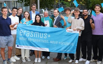 Schulklasse mit Banner mit dem Wortlaut Erasmus. Zusätzlich hat jeder noch eine kleine blaue Erasmus Fahne. Die Gruppe steht draußen, neben der Schule