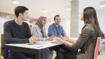 Bild von Studierenden der Wirtschaftsuniversität Wien.  Zwei Männer und zwei Frauen sitzen um einen eckigen Tisch und unterhalten sich. Eine Frau trägt ein fliederfarbiges Kopftuch, alle Studierenden lächeln.
