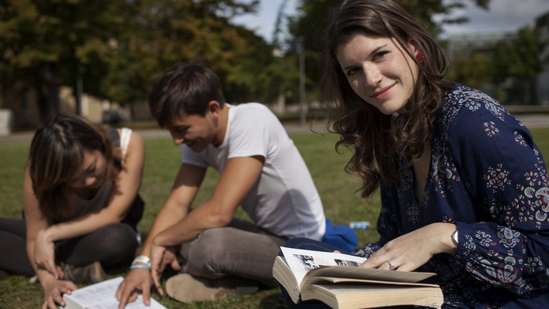 Gruppe sitzt auf Wiese und sieht in Bücher, junge Frau lächelt in die Kamera