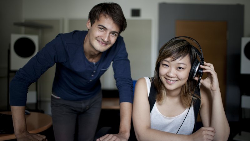 Ein Student und eine Studentin an einem Mischpult mt Kopfhörern lachen in die Kamera