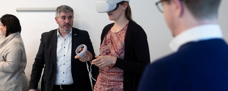 Eine Frau trägt eine VR-Brille und bedient eine VR-Steuerung. Zwei Männer schauen ihr dabei zu.