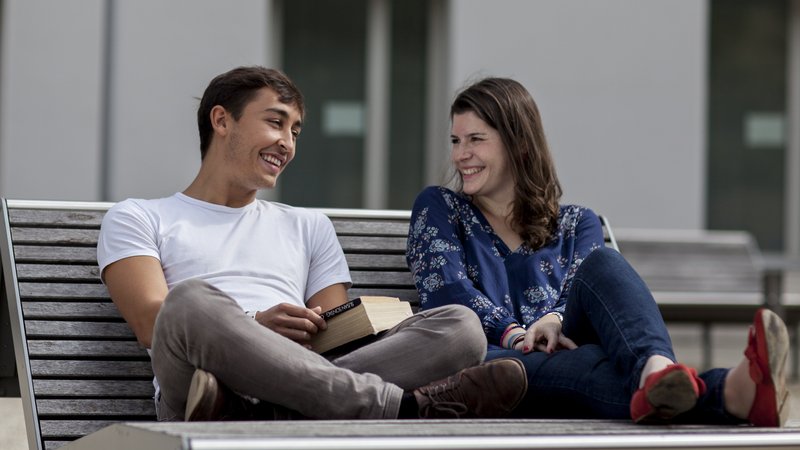 Junger Mann und junge Frau sitzen auf Bank vor Universität